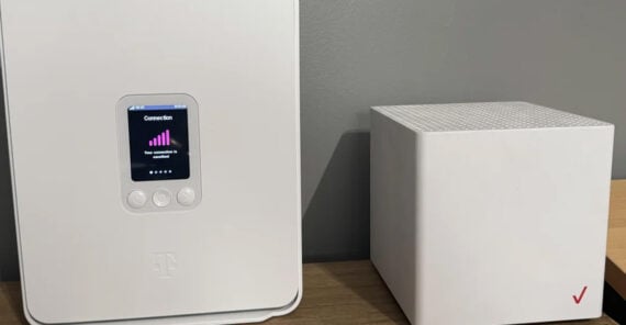 A T-Mobile 5G Home Internet gateway with a small screen showing signal strength sits next to a white Verizon router on a wooden surface against a gray wall.