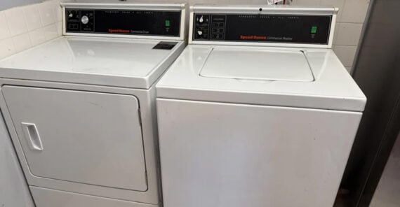A white commercial dryer and washing machine sit side by side in a laundry room. Both have control panels with buttons and coin slots above the front-loading doors.