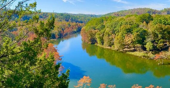 A river winds through a lush, forested landscape with trees showing green and hints of autumn colors under a clear blue sky.