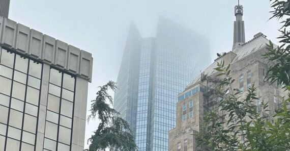 Tall modern skyscraper with a glass exterior partially obscured by fog, surrounded by older stone buildings and green trees in the foreground.