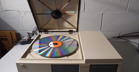 A close-up of a vintage LaserDisc player with its lid open, showing a large reflective LaserDisc inside, set against a gray concrete wall background.