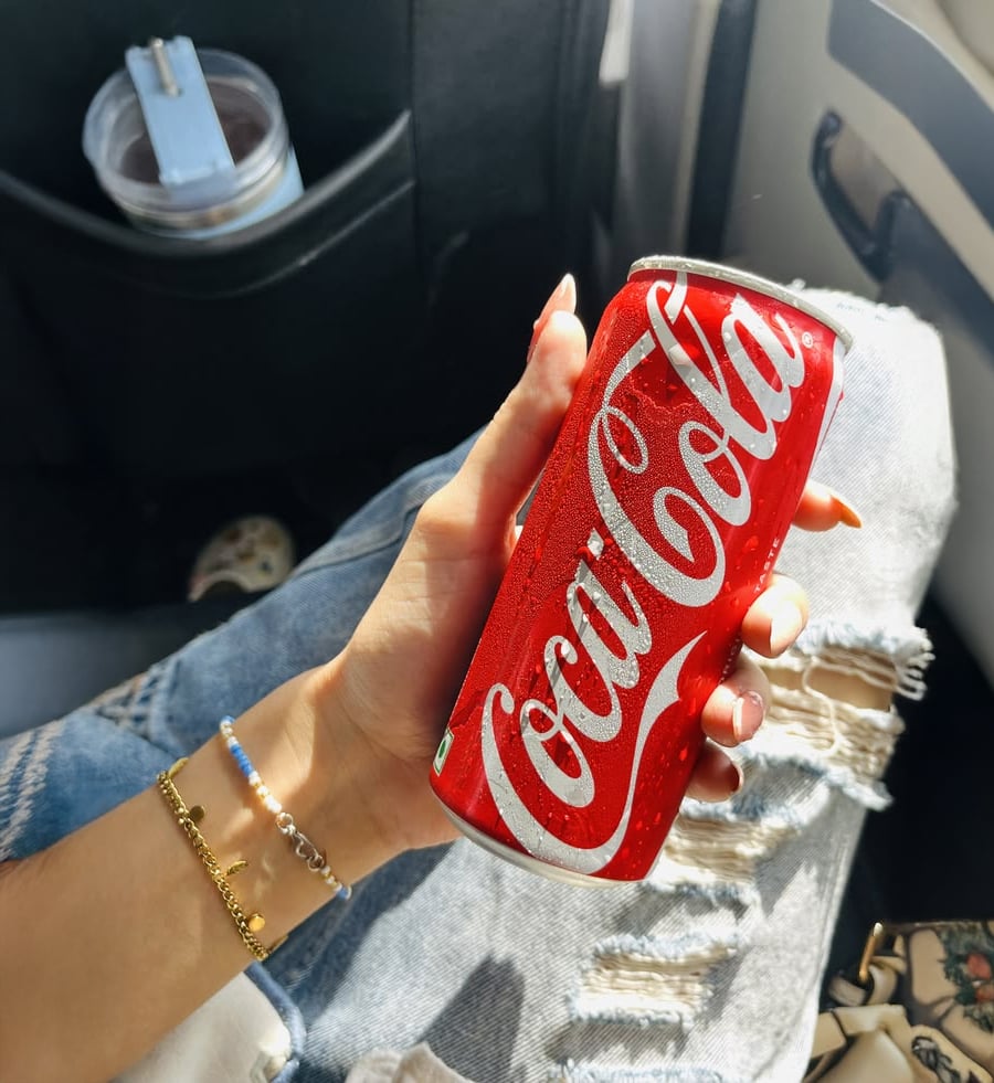 A person wearing ripped jeans and a bracelet holds a cold, red Coca-Cola can inside a car, with sunlight shining on their hand.