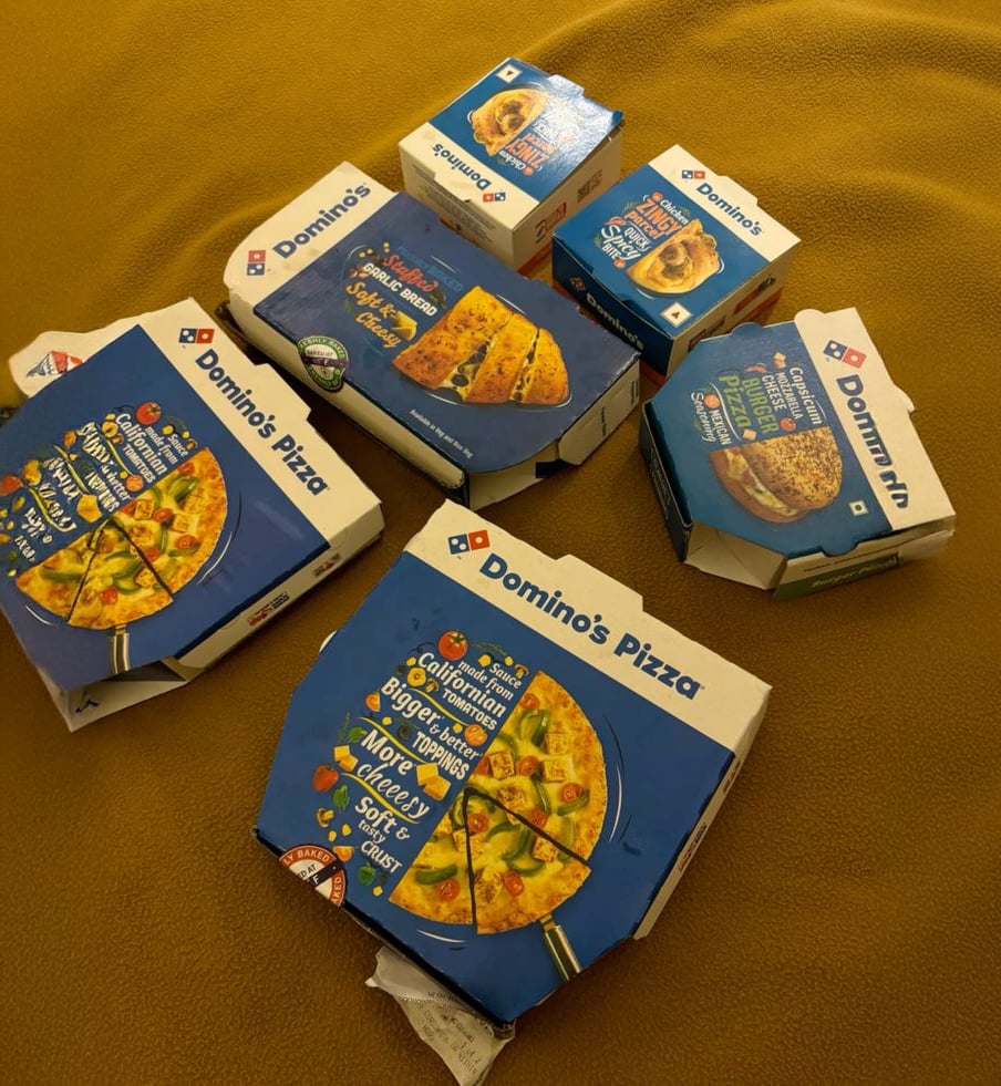 Several closed Domino’s Pizza boxes and dessert containers are arranged on a mustard yellow blanket. The boxes are various sizes for pizza, breadsticks, and desserts.