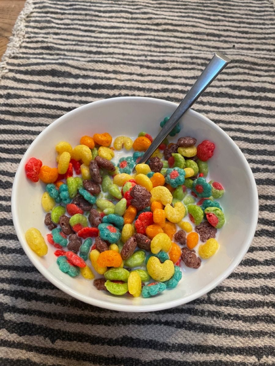 A white bowl filled with colorful, fruit-shaped cereal and milk sits on a black-and-white striped cloth. A metal spoon rests inside the bowl.