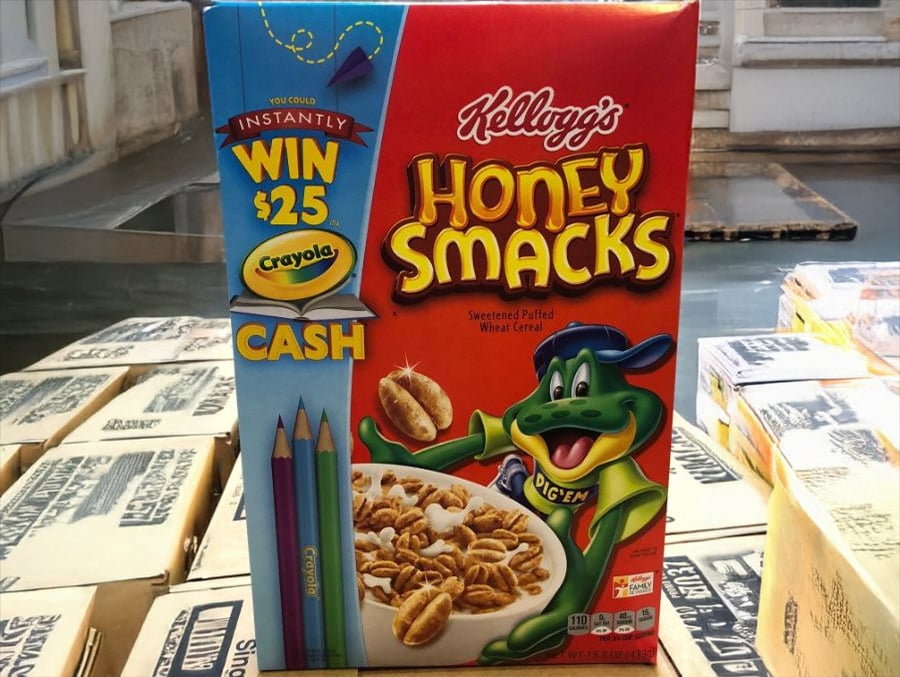 A box of Kellogg’s Honey Smacks cereal featuring a frog mascot, a bowl of cereal, crayons, and a promotion to win $25 Crayola cash. The box is on top of stacked cardboard boxes.