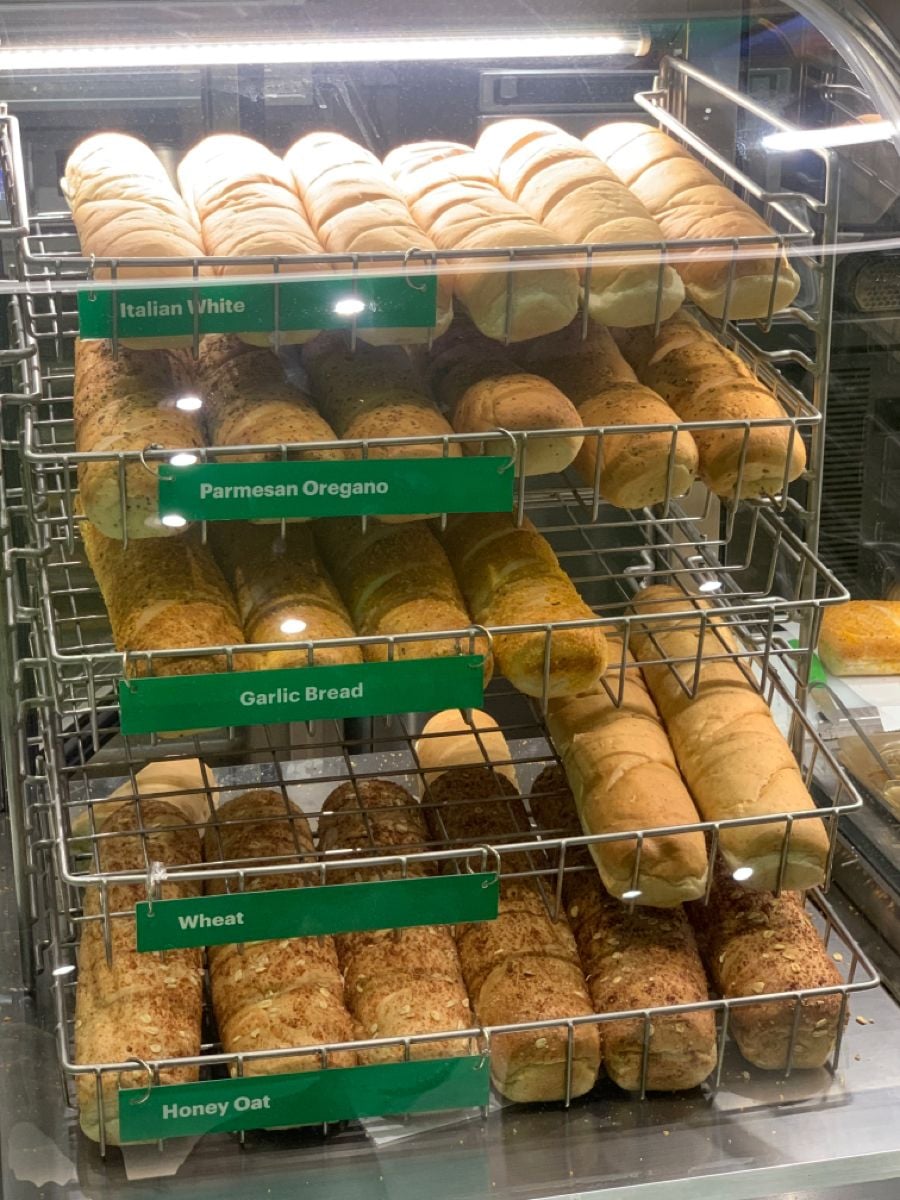 A bakery display case with shelves holding loaves of bread labeled: Italian White, Parmesan Oregano, Garlic Bread, Wheat, and Honey Oat. Each type is organized in separate rows with green signs.