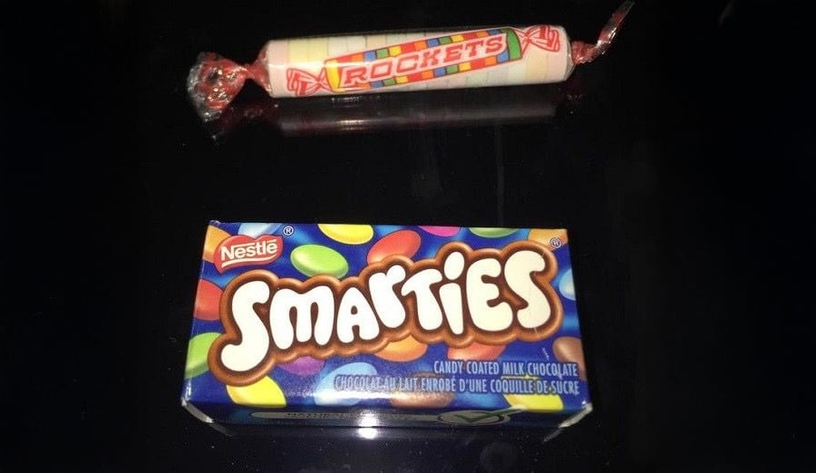A roll of Rockets candy and a box of Nestlé Smarties are placed on a black surface. The Rockets are wrapped in white paper, and the Smarties box is blue with colorful candy images.