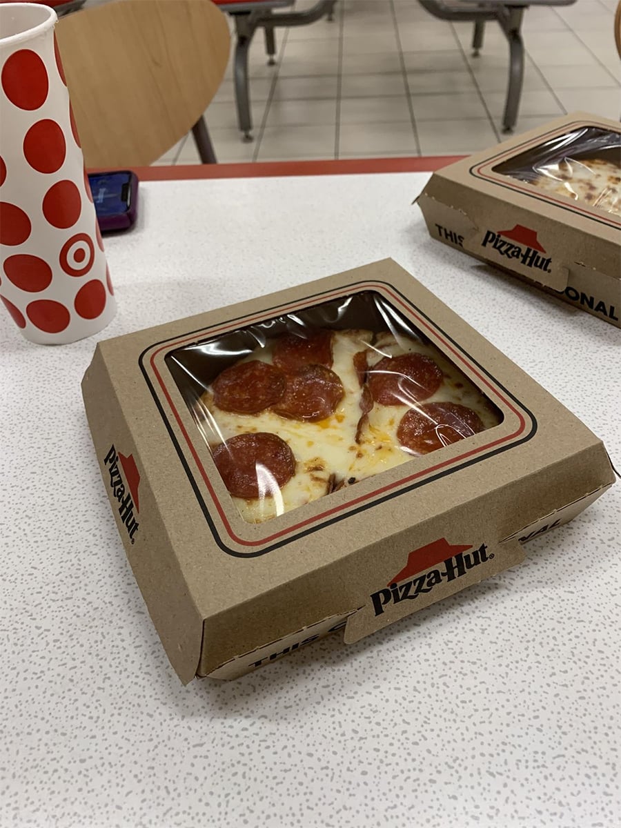 A Pizza Hut box with two slices of pepperoni pizza sits on a white table next to a red Target cup. Another pizza box and empty chairs are visible in the background.