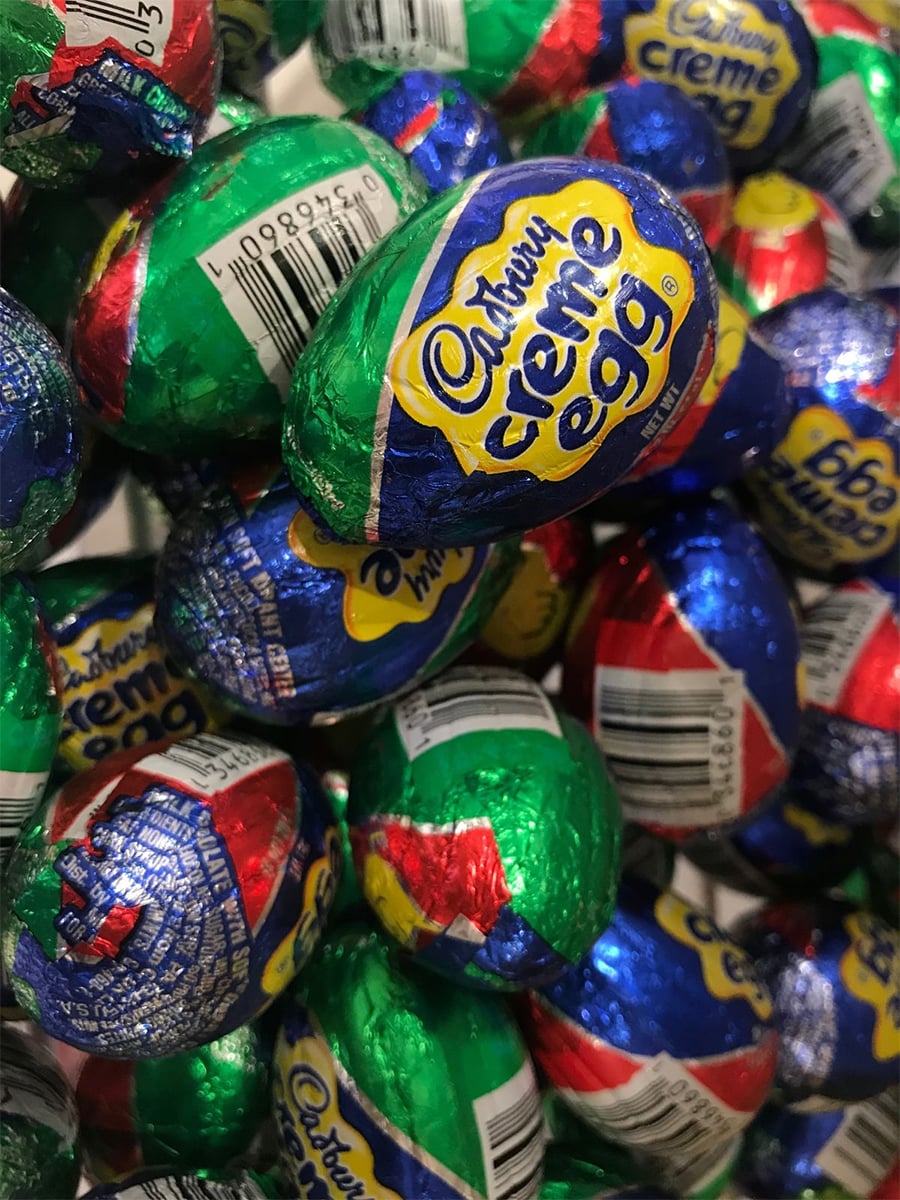 A pile of foil-wrapped Cadbury Creme Eggs with colorful blue, red, green, and silver packaging, featuring the yellow Cadbury logo and barcode stickers visible on some eggs.