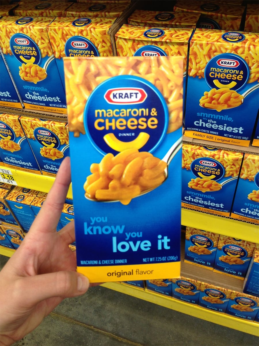 A hand holds a blue box of Kraft Macaroni & Cheese Dinner, original flavor, in front of a store shelf filled with more boxes of the same product.
