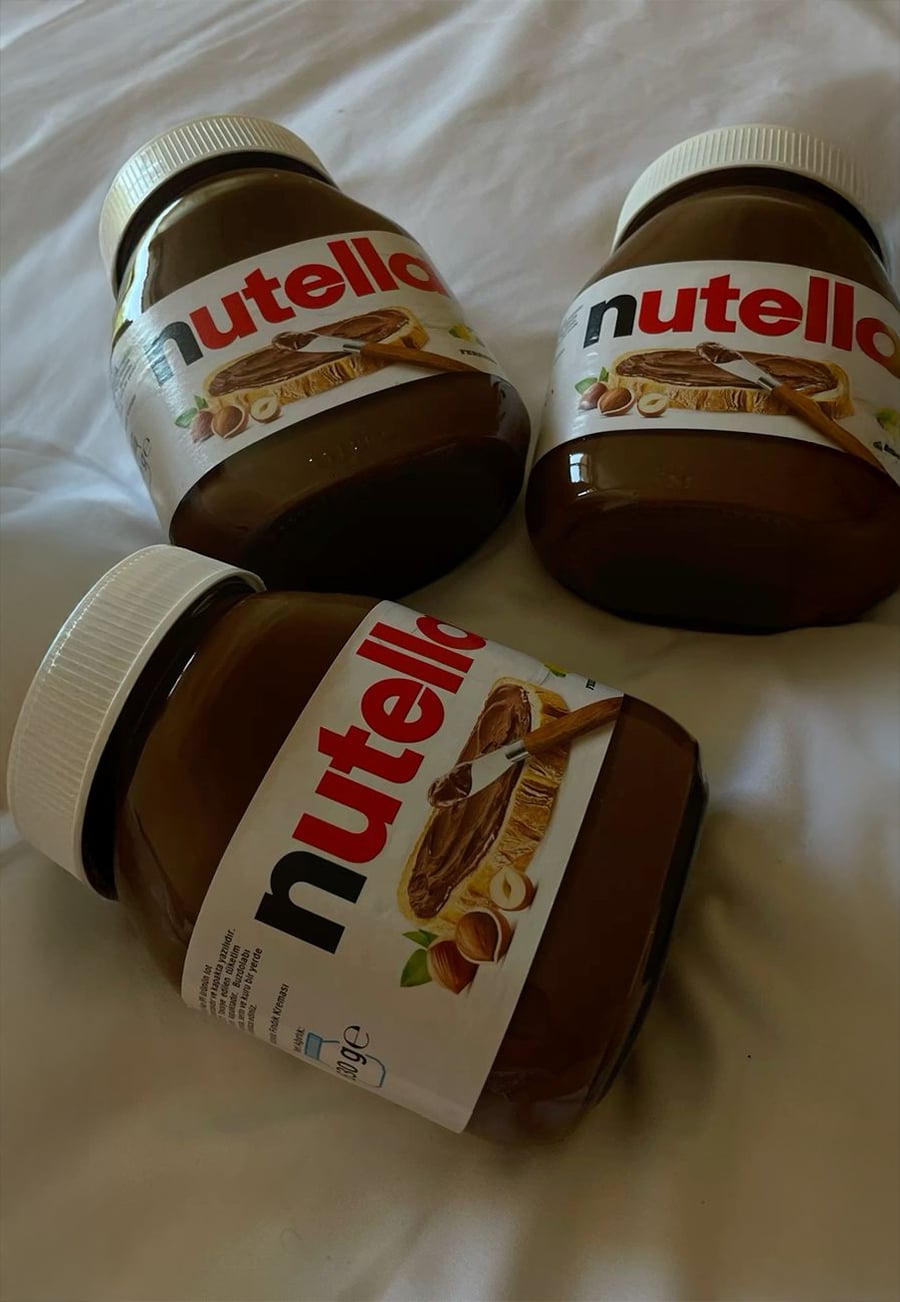 Three jars of Nutella, with white lids and labeled sides, are lying on a white surface. The jars are arranged at different angles, with two upright and one lying sideways in the foreground.