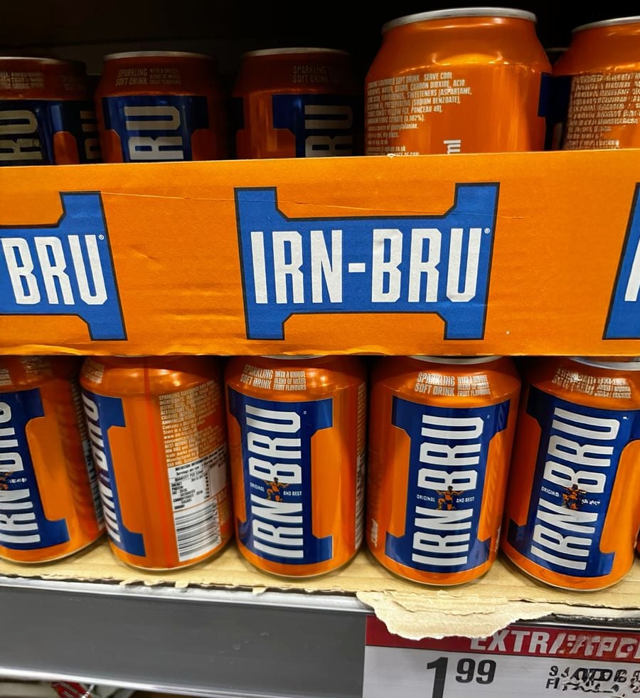 Cans of IRN-BRU with bright orange packaging are stacked on a shelf, with some visible price tags below them.