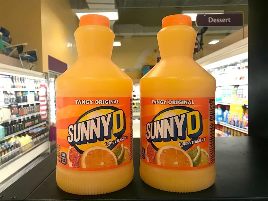 Two bottles of SunnyD Tangy Original orange drink are displayed on a shelf in a grocery store, with coolers and a “Dessert” aisle sign visible in the background.