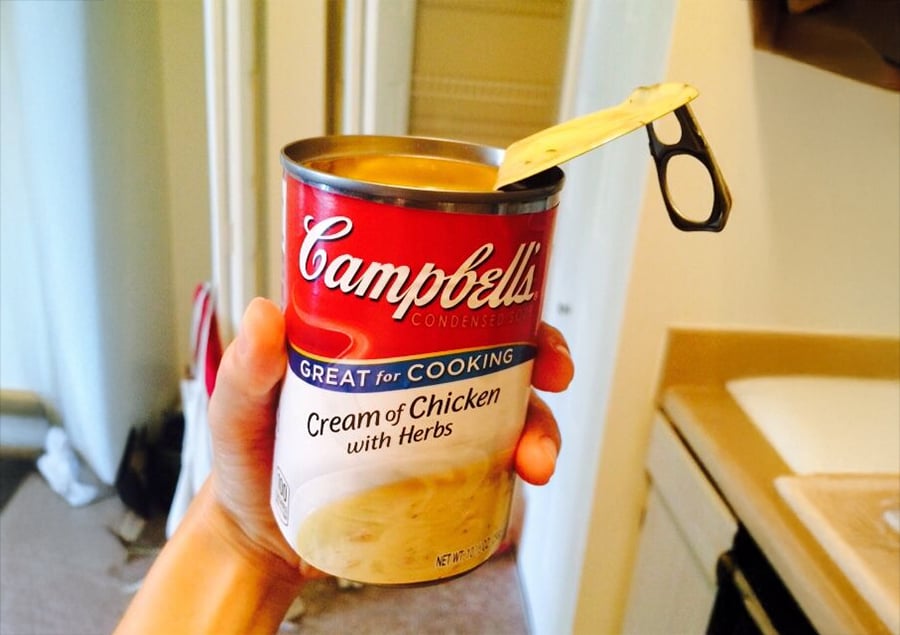 A hand holds an open can of Campbell’s Cream of Chicken with Herbs soup in a kitchen, with the lid still attached.