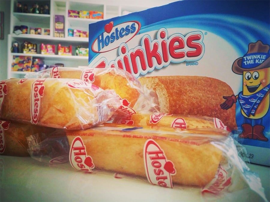 Individually wrapped Hostess Twinkies are stacked in front of a blue Twinkies box, with shelves of colorful toys and collectibles blurred in the background.