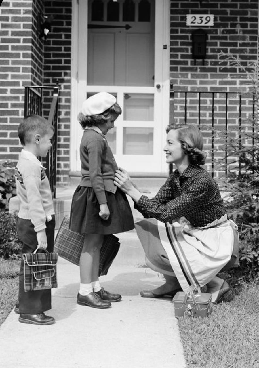 A woman kneels to help a young girl with her coat as a boy stands nearby. Both children hold lunchboxes, suggesting they’re on their way to school. They are outside a brick house with the number 239 by the door.
