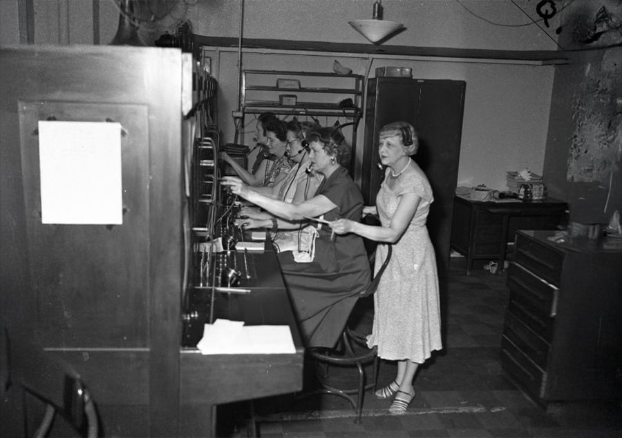 Four women operate a vintage telephone switchboard in a black-and-white photo; one stands while the others sit, all focused on connecting calls.