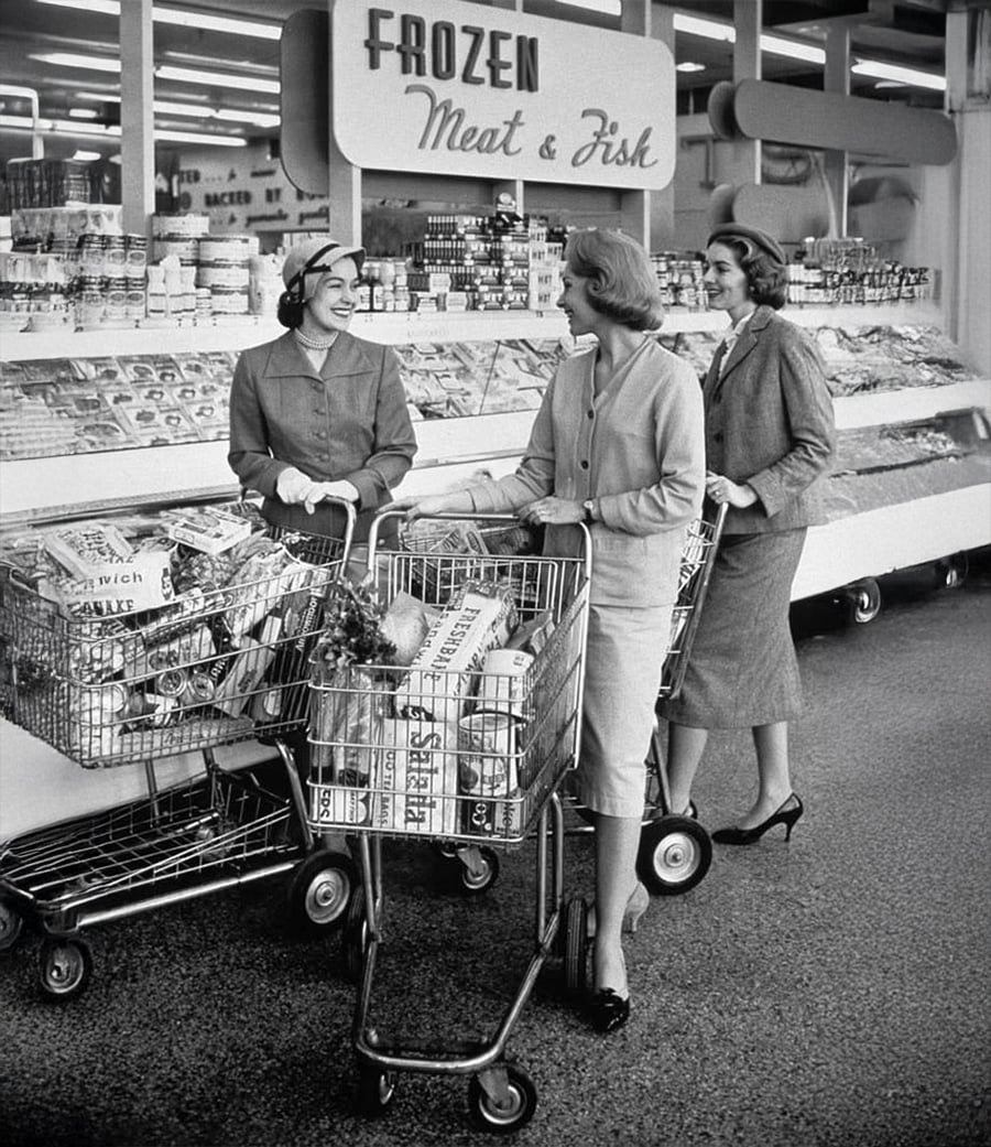 Three women in 1950s-style clothing smile and chat while shopping in a supermarket aisle labeled “Frozen Meat & Fish.” Each woman has a grocery cart filled with food items.