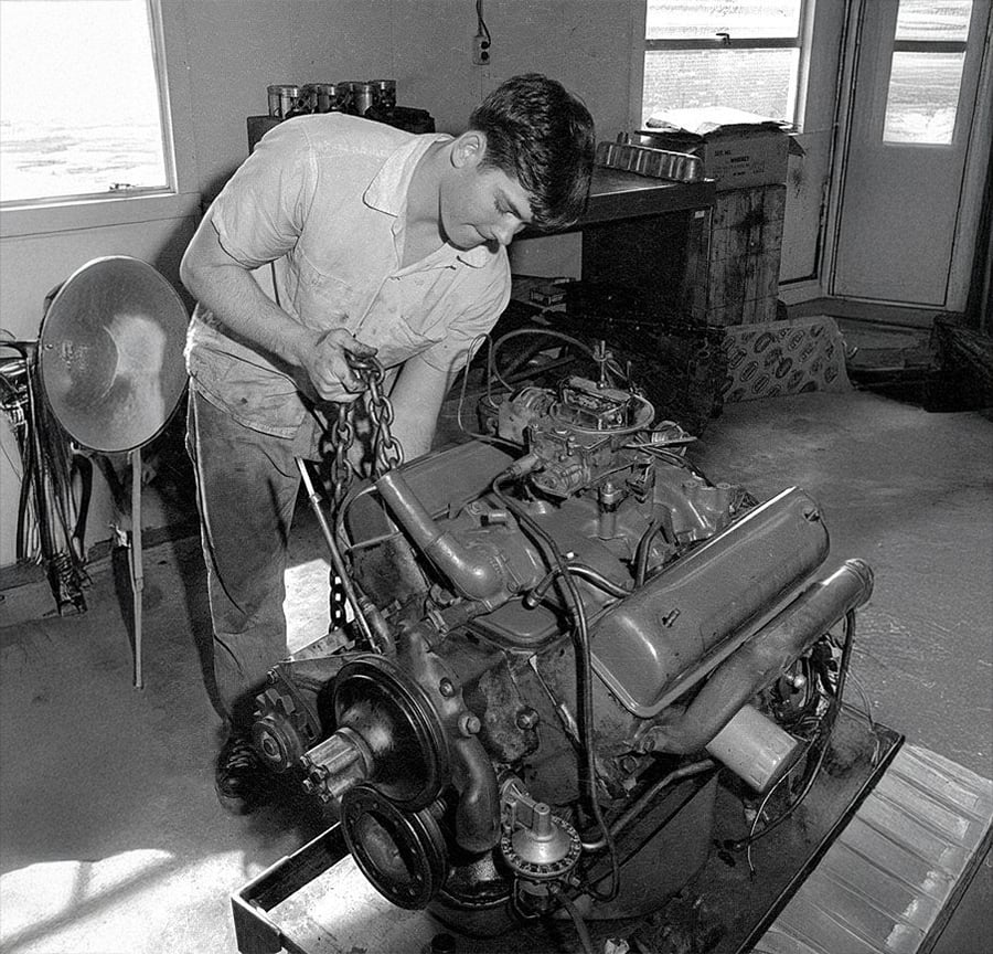A man in a workshop uses a chain hoist to lift a large car engine. Tools and equipment are visible around him, and light comes in through the windows and door in the background.