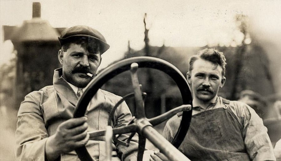 Two men with mustaches sit behind the wheel of an old vehicle. The man on the left wears a cap and smokes a cigarette, while the man on the right looks at the camera. Both wear work clothes. The image is in black and white.