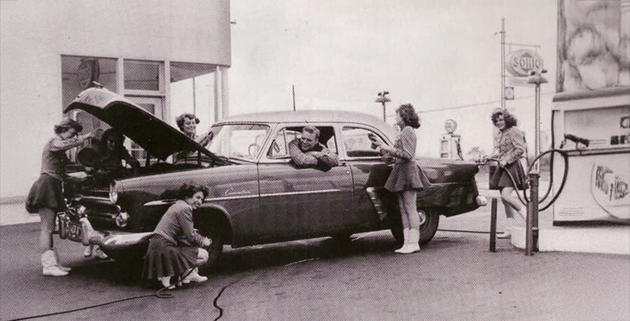 Five women in 1950s-style outfits pose around a classic car at a gas station, some pretending to work on the engine while a man sits inside the car smiling. A gas pump and Sohio sign are visible in the background.