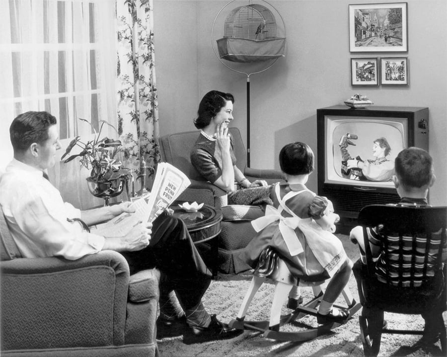 A 1950s family sits in a living room watching TV. The father reads a newspaper, the mother smiles, and two children watch the show. The room has vintage decor and a birdcage.