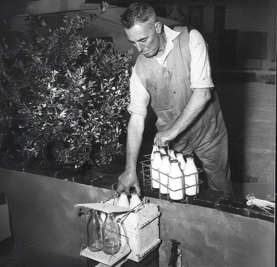 A man in work clothes delivers glass bottles of milk, placing them into a box on a porch. He holds a metal carrier with more bottles. There is a plant nearby and the scene appears to be from an earlier era.