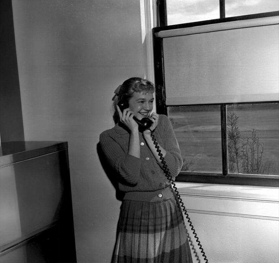 A woman in a plaid skirt and cardigan smiles while talking on a corded telephone near a window with a view outside.