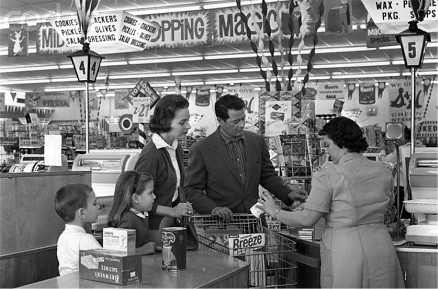 A family with two children stands at a grocery store checkout counter while a cashier rings up their items. The store is decorated with sale signs and banners overhead.
