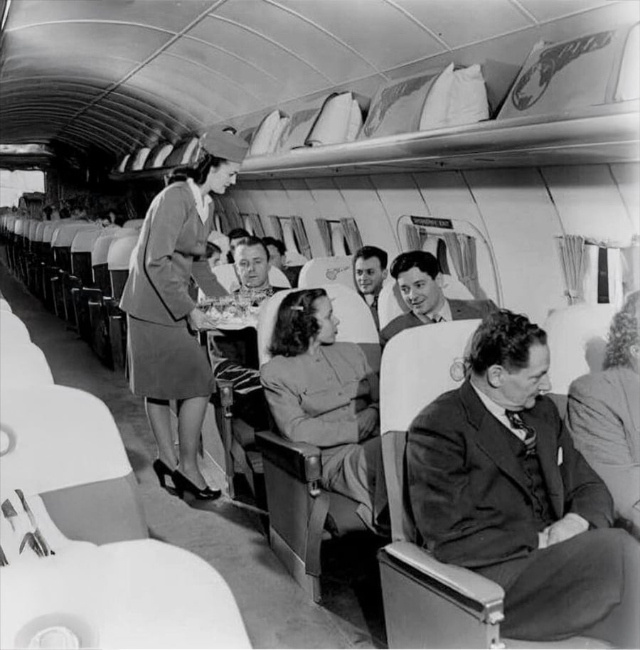 A flight attendant serves drinks to passengers seated in a vintage airplane cabin. The passengers are dressed in formal attire, and overhead compartments hold luggage. The scene appears to be from the mid-20th century.