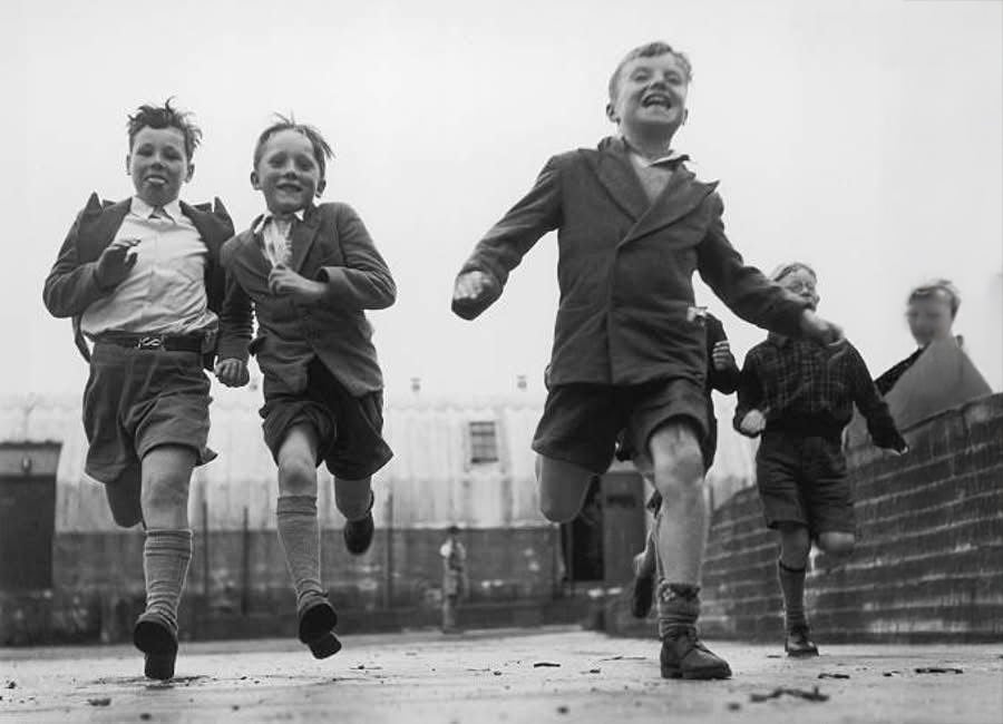 Four young boys in vintage clothing run joyfully toward the camera on a paved street, with blurred buildings and another child in the background under a cloudy sky.