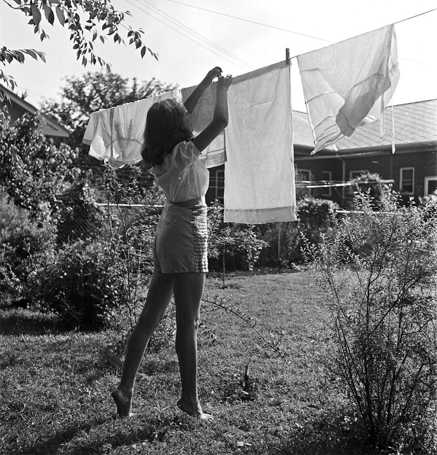 A woman stands barefoot on grass, reaching up to hang laundry on a clothesline in a sunny backyard, with houses and trees in the background.