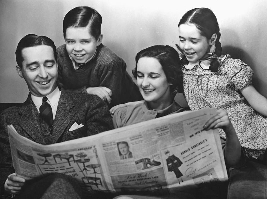 A smiling family of four sits together. The father holds a newspaper, the mother sits beside him, and two children, a boy and a girl, look happily at the newspaper over their parents’ shoulders.