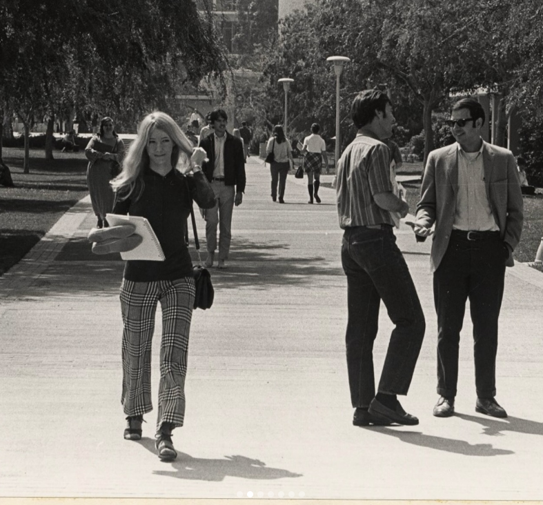 Black-and-white photo of students walking on a campus path. A woman with long hair and plaid pants holds papers, while two men stand talking nearby. Trees and more students are visible in the background.