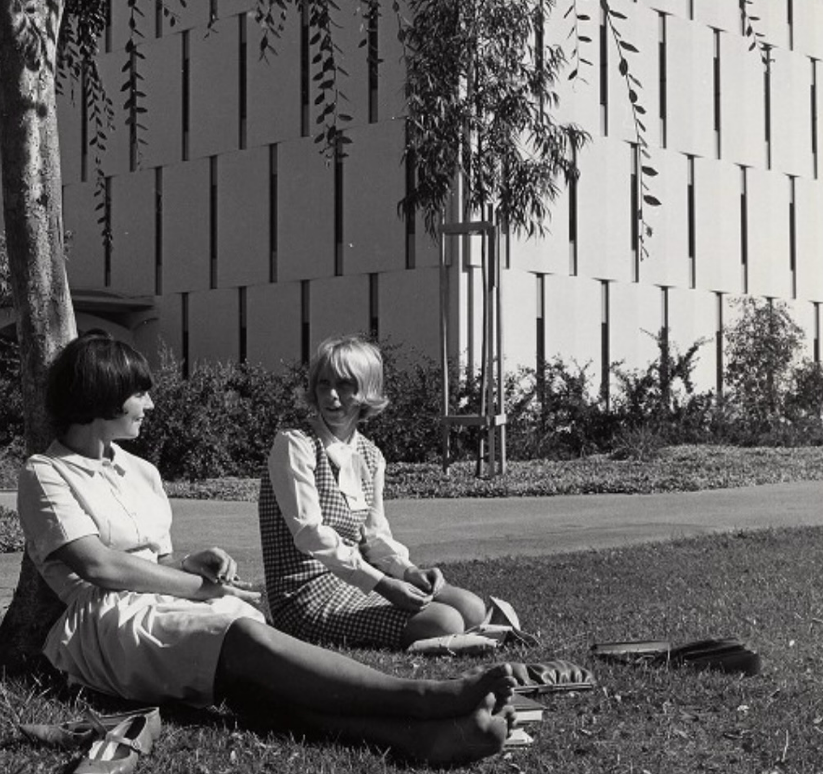 Two women sit on grass in the shade of a tree, talking and relaxing barefoot. Behind them is a modern building with geometric patterns and small trees, suggesting a campus setting.