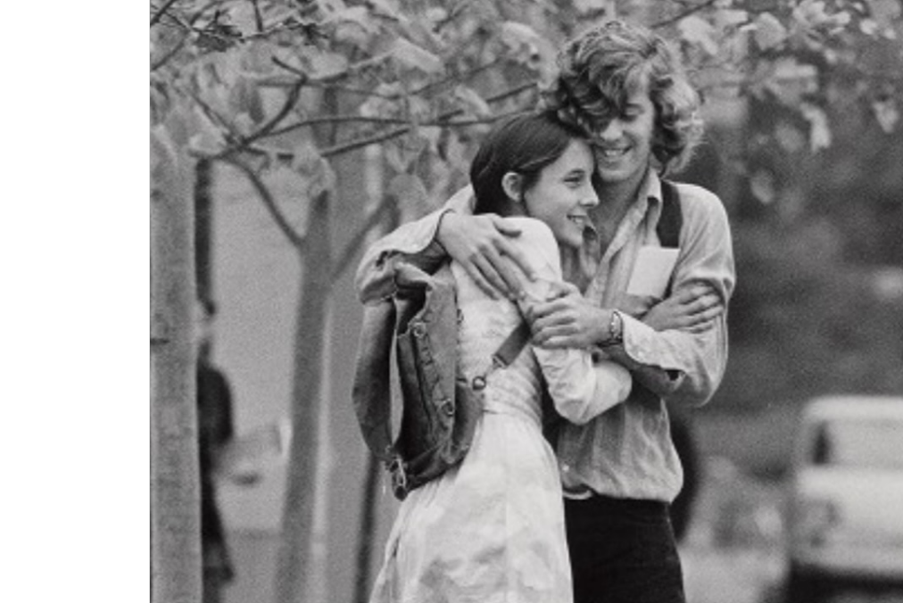 A young man and woman embrace and smile under trees. The woman wears a light dress and backpack; the man hugs her from behind, wearing a casual shirt. The photo is black and white, capturing a candid, joyful moment.