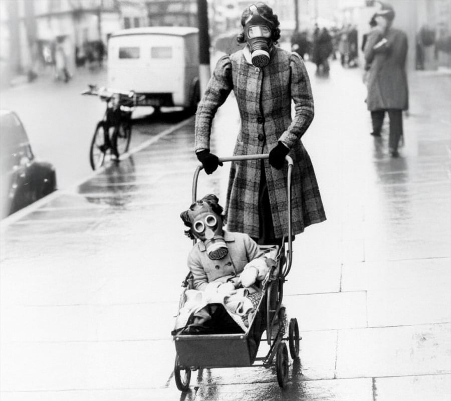 A woman and a child, both wearing gas masks, walk outside. The woman pushes the child in a stroller on a wet city sidewalk. The scene appears to be from the past, possibly during wartime.