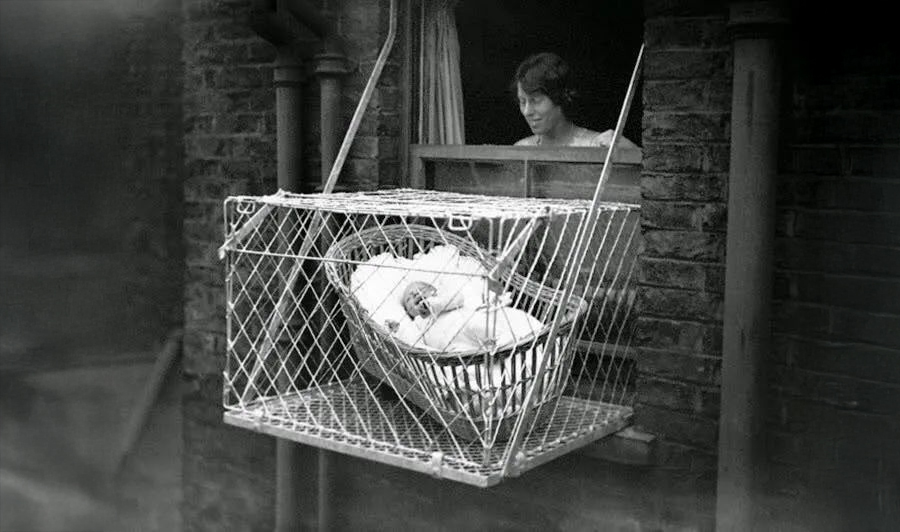 A woman looks out a window at a metal cage containing a baby in blankets, suspended outside a brick building, likely for fresh air.
