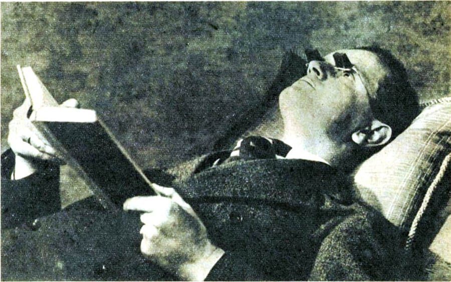 A person wearing glasses lies on a pillow, holding and reading a book above them while resting on their back, dressed in a suit and tie.