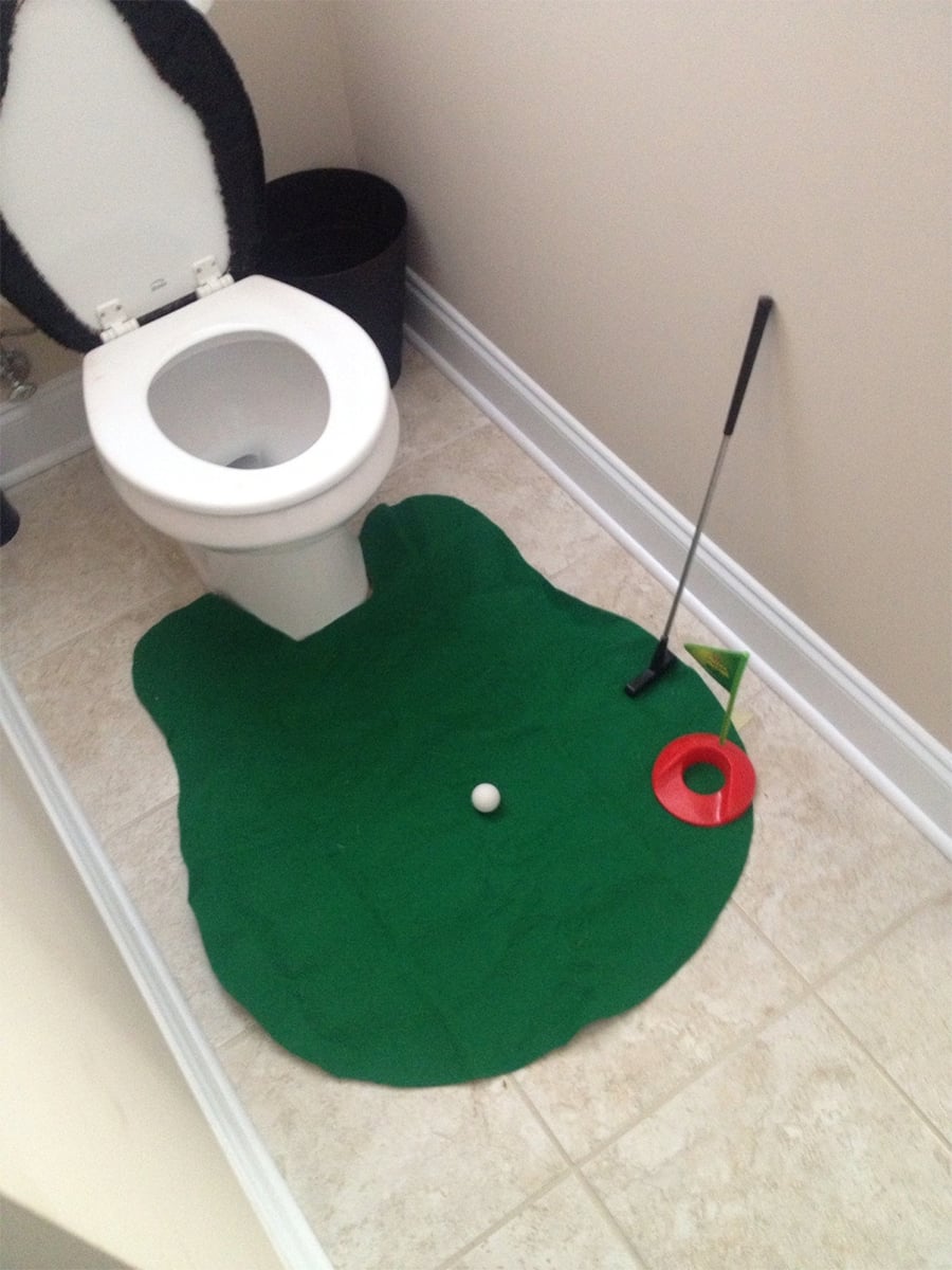 A toilet with a green putting mat, golf ball, putter, and hole set up on the bathroom floor, resembling a mini-golf course.