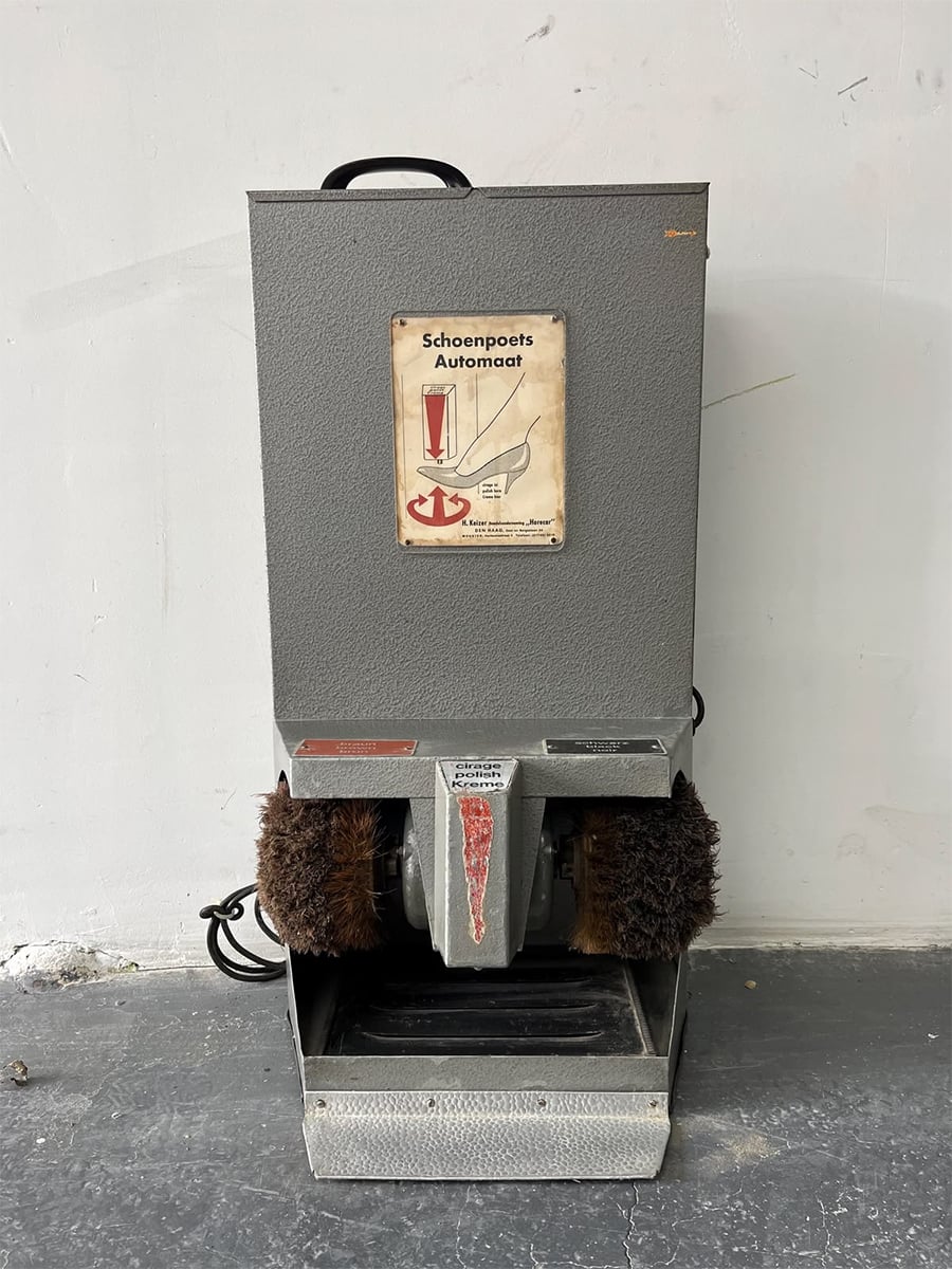 A vintage gray shoeshine machine with two brown brushes, a metal base, and a sign labeled “Schoenpoets Automaat” with a shoe illustration, standing against a white wall on a concrete floor.