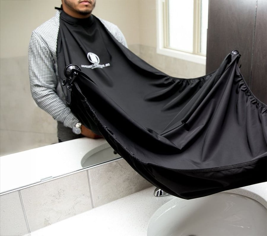 A person wearing a black grooming cape attached to a mirror, creating a barrier above the sink to catch hair trimmings during shaving or haircutting.