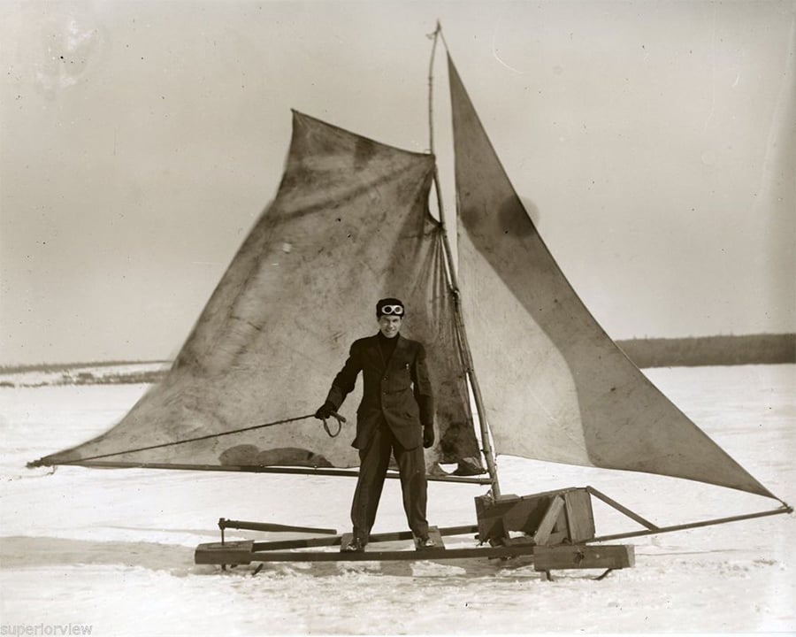 A person in a suit and goggles stands on a vintage ice sailboat with large triangular sails on a frozen, snowy lake.