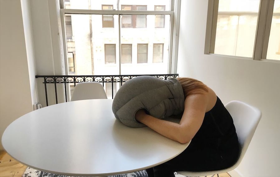 A person sits at a round table with their head inside a gray pillow designed for napping, in a bright room with large windows.
