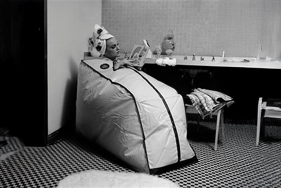 A person sits in a portable sauna bag, head exposed, wearing a towel on their head and reading a book in a tiled room with a long counter and chair.