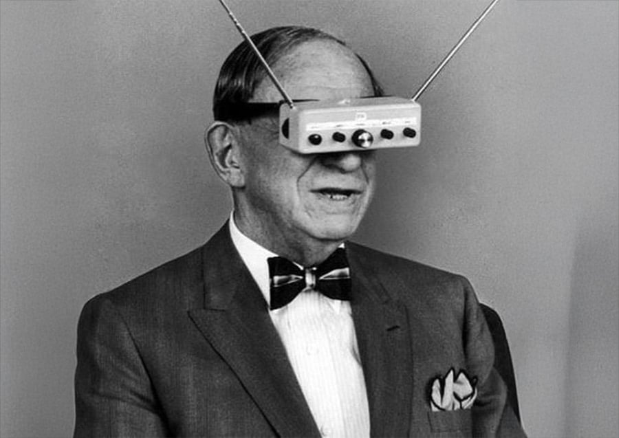 An older man in a suit and bow tie wears a device with antennas over his eyes, resembling early virtual reality or television glasses, against a plain background.