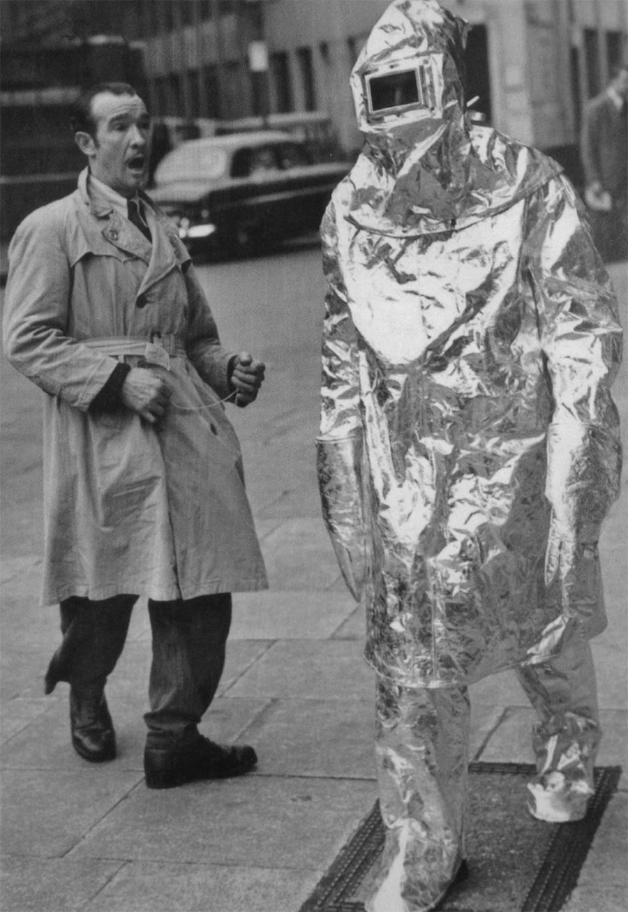 A person in a shiny, metallic hazmat suit walks on a city sidewalk while another person in a trench coat looks on with a surprised expression.