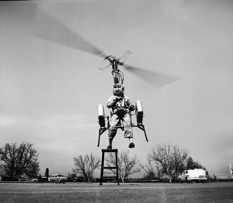 A person tests a small personal helicopter device while hovering above a platform outdoors, with blurred spinning blades and trees and cars in the background.