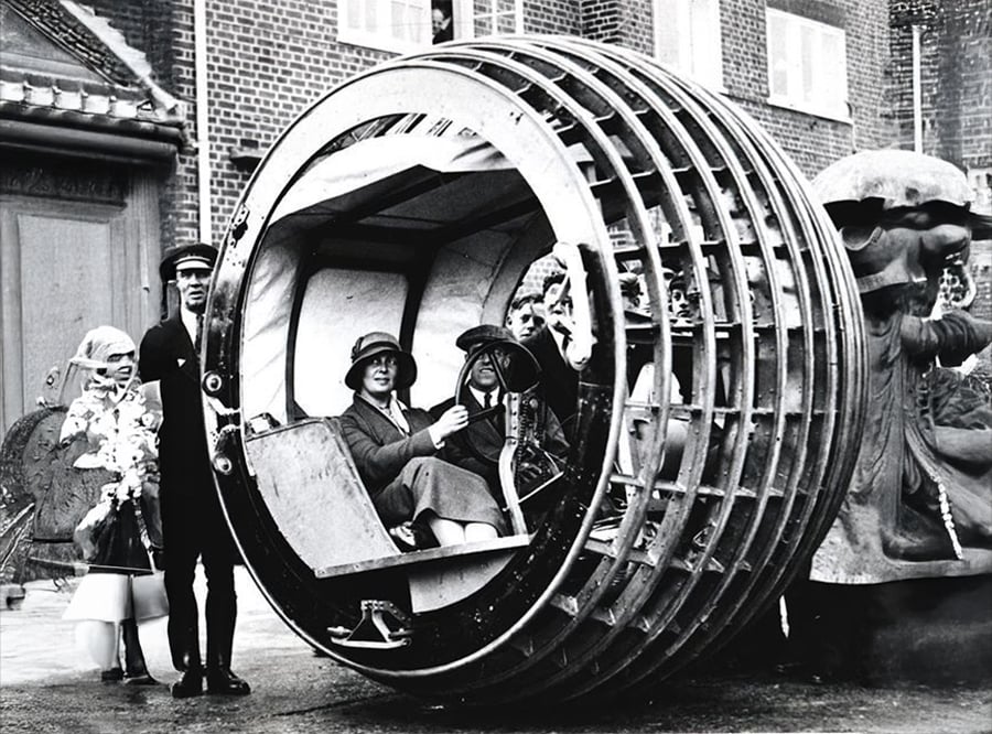 Four people sit inside a large, circular, wheel-like vehicle, while a man in uniform stands beside it. The scene appears vintage, with brick buildings and an ornate statue in the background.