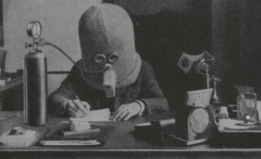 A person sits at a desk writing while wearing a large, helmet-like mask covering their entire head, connected to a tank. Old-fashioned office equipment and papers are on the desk.